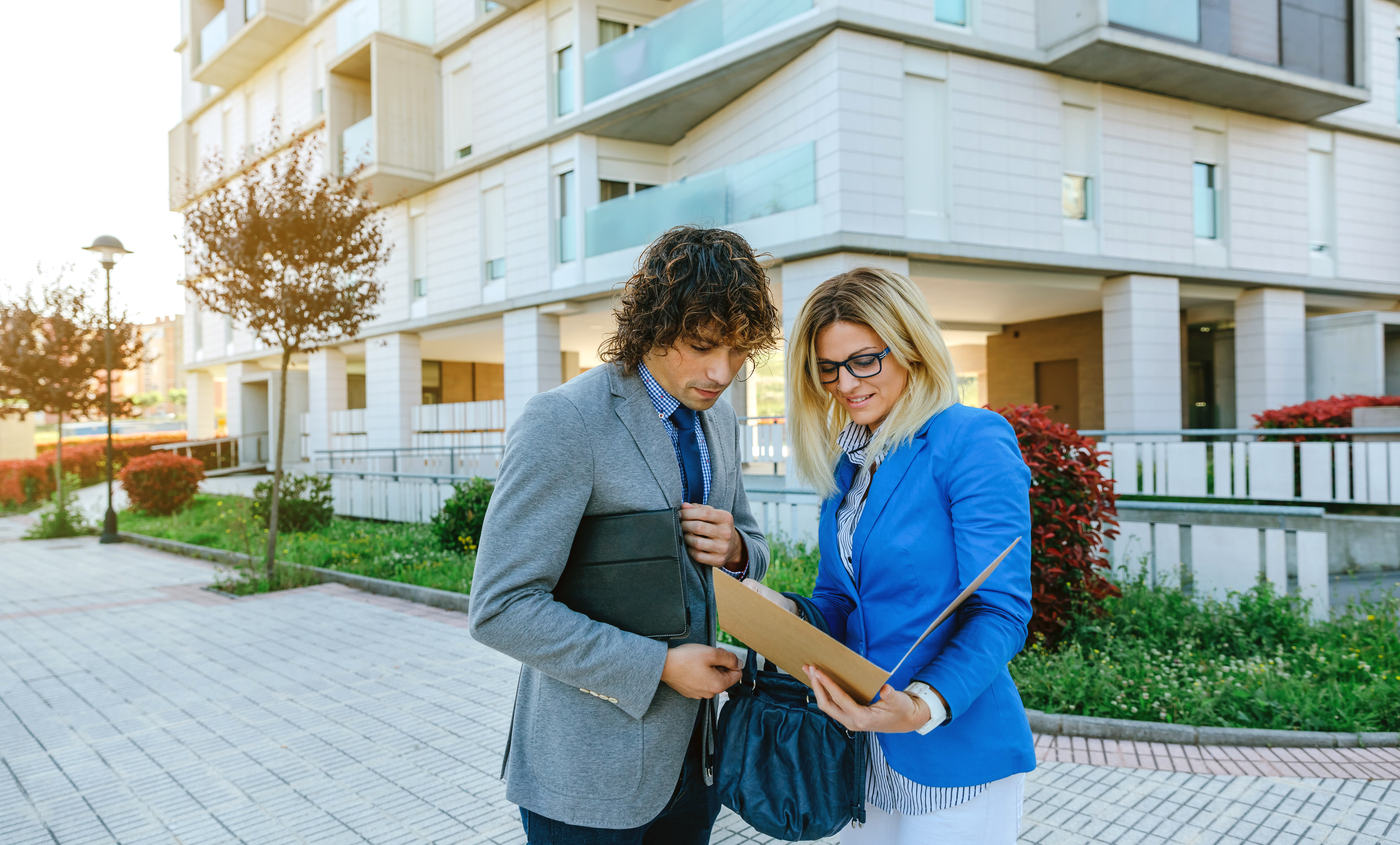Property manager and owner reviewing documents in a folder outside a modern apartment building with balconies and landscaping